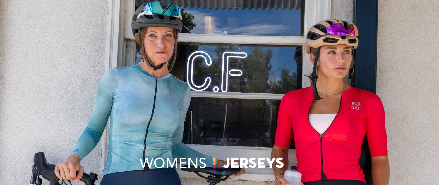 Two female cyclists in a blue and red jesery outside a coffee shop holding a bike. 